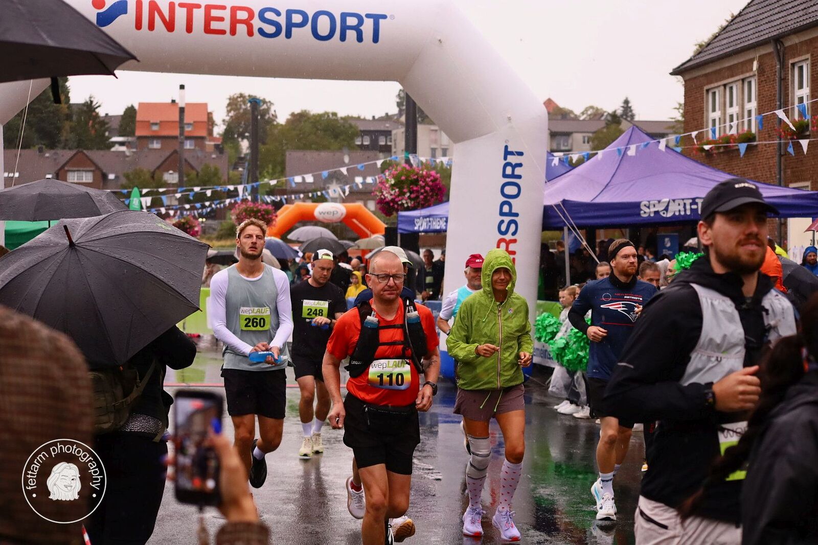 Läuferinnen und Läufer passieren im Regen den Startbogen, Zuschauer mit Regenschirmen am Rand
